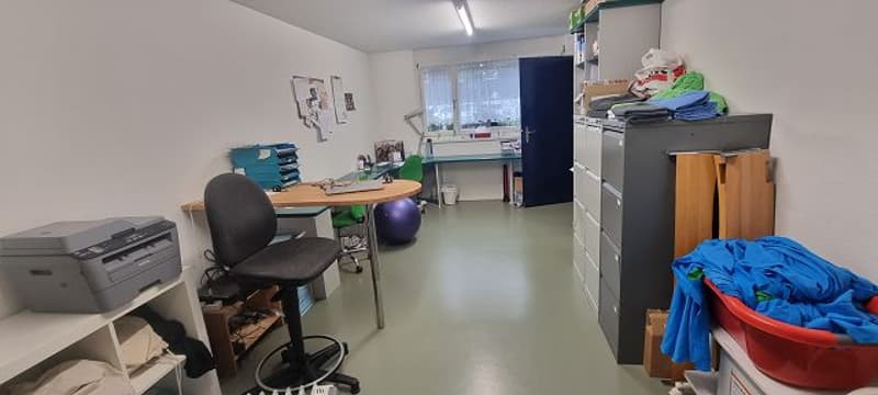 The image shows a small office or workspace. It has a desk with a computer, a printer, and other office equipment. There is also a washing machine visible, indicating that this space may be used for both work and personal activities.