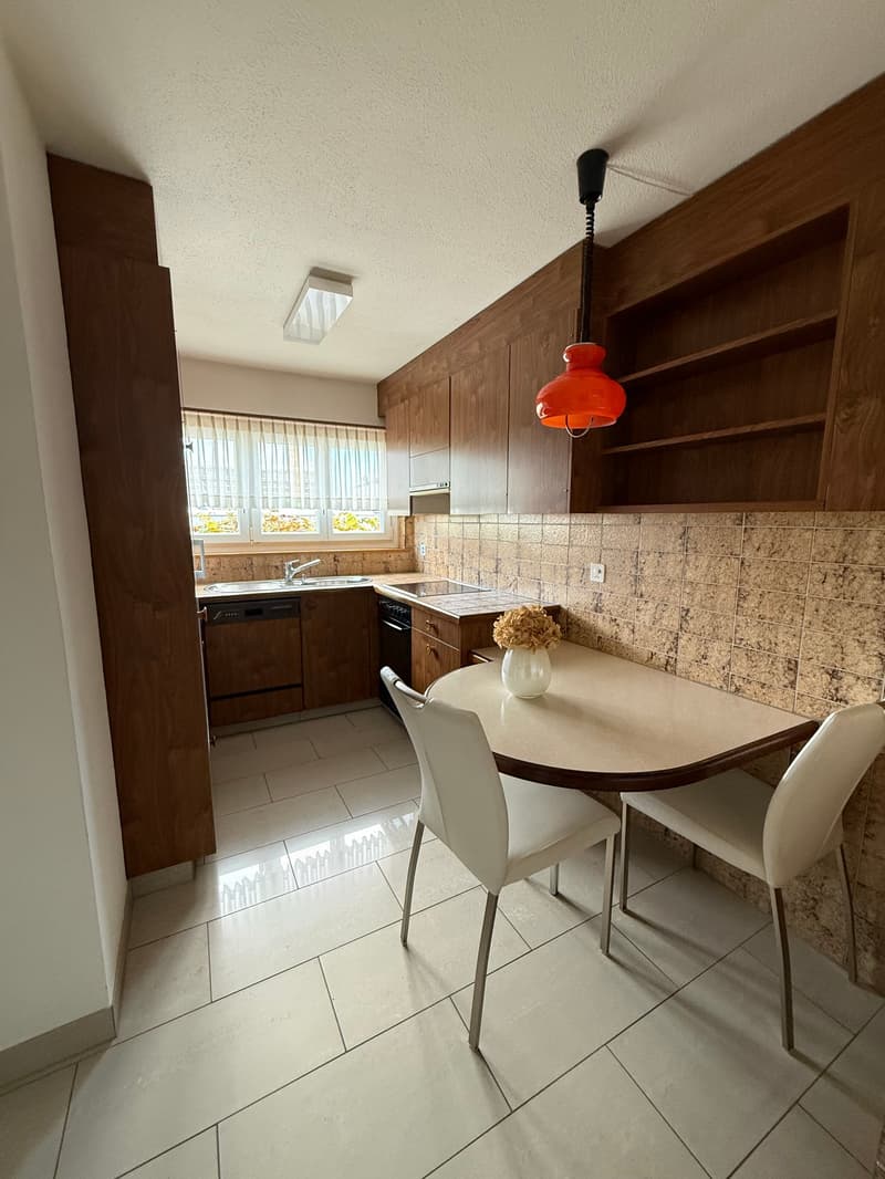 Kitchen with white tiled flooring, modern white chairs around a round dining table, wooden cabinets, stainless steel kitchen appliances, and natural light from a window.