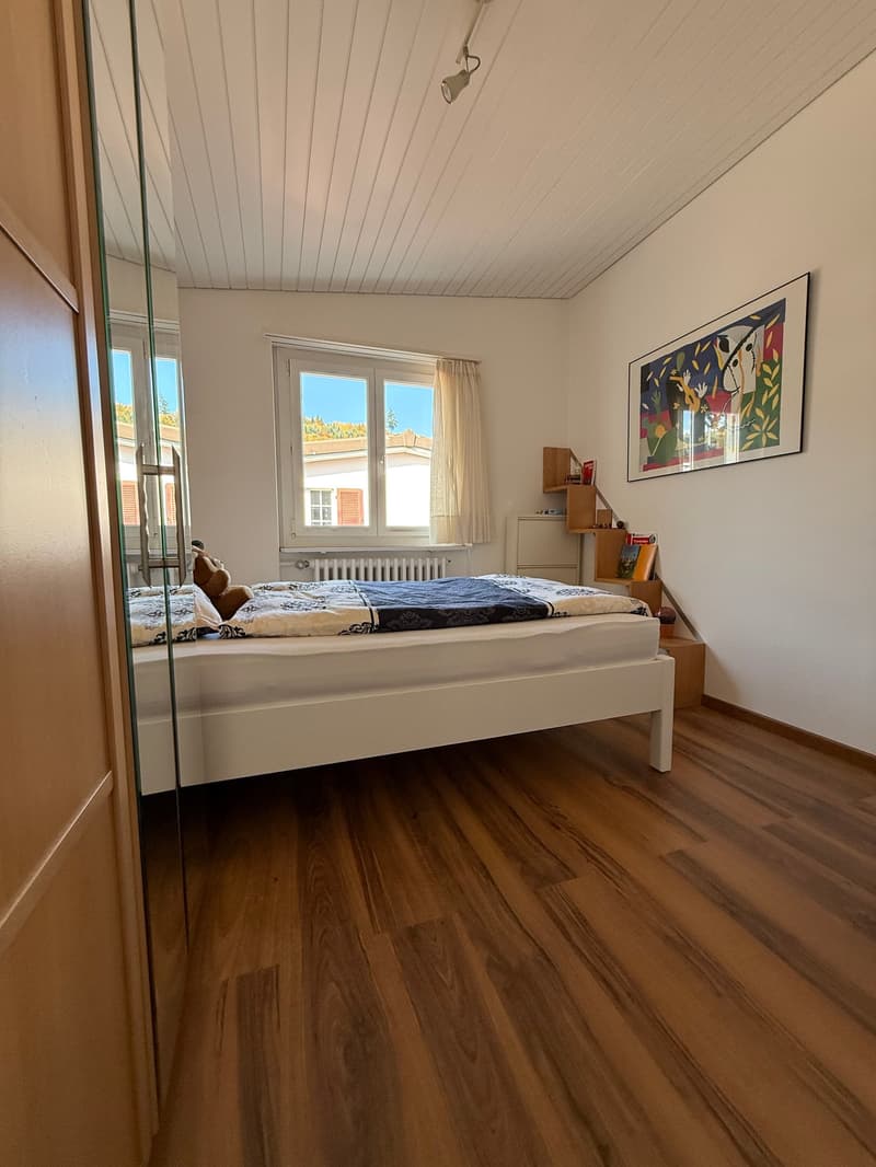 Bedroom: hardwood floor, white walls, a white bed with a decorative blue comforter, bed headboard, white nightstands on both sides, a shelf mounted on the wall near the window, a framed picture on the wall, curtains, a small stuffed animal on the bed.