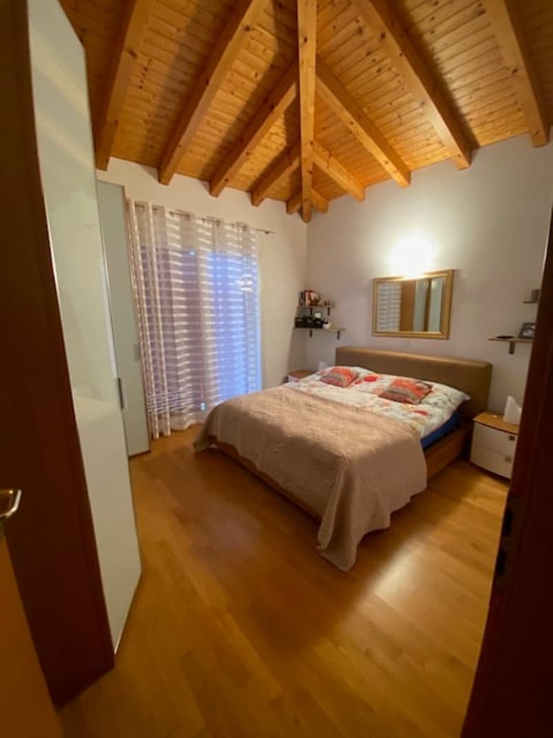 Bedroom with wooden floor, wooden beams on ceiling, white walls, large bed, wooden nightstand, mirror on wall, white curtains