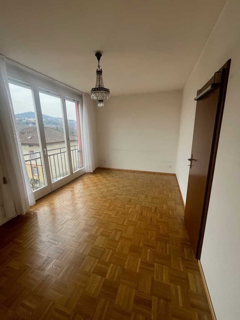 Spacious room with large windows, hardwood parquet flooring, and a chandelier light fixture. The room has a balcony or terrace accessible through the windows.