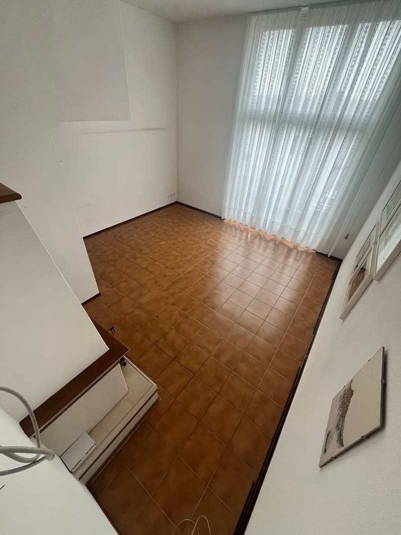 An empty room with wooden flooring, white walls, and a large window with white curtains. There are picture frames hanging on the right wall.