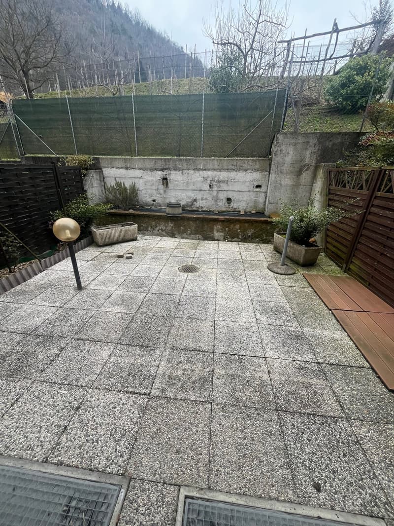 Paved patio area with concrete tiles, surrounded by a wire fence and vegetation. There appears to be a small pond or water feature in the center of the space.