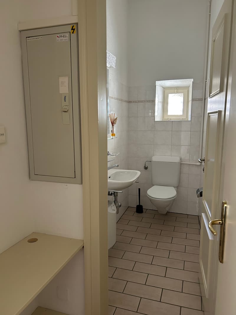 Small bathroom with white tiled walls, white sink, white toilet, shower window, bench, and a trash bin.