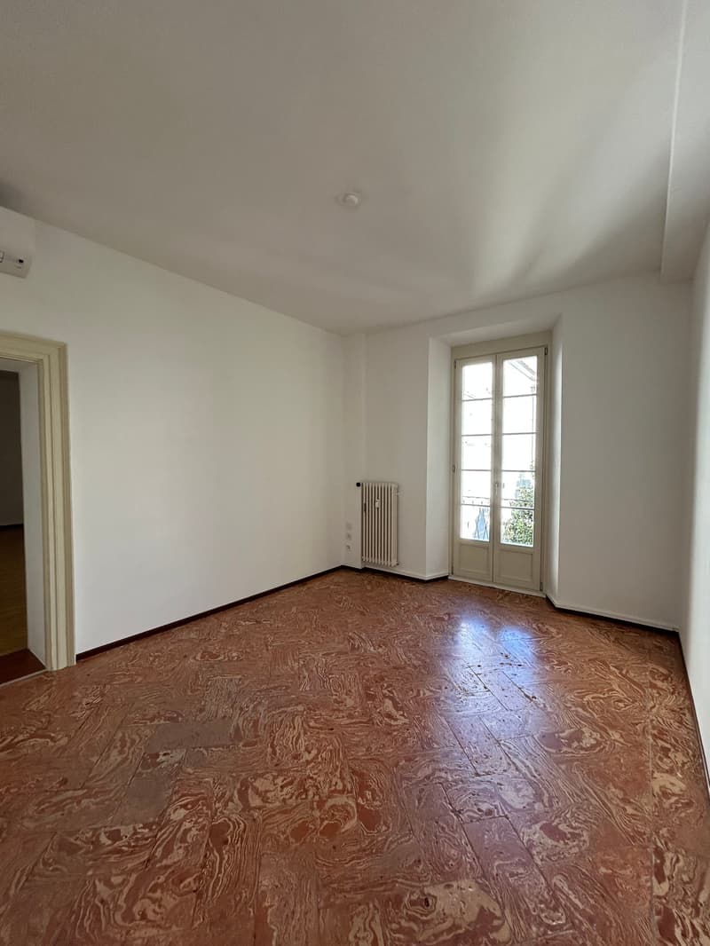 This image shows an empty room with white walls and a wooden floor. The room has a window that lets in natural light, and there is a radiator visible on one of the walls.