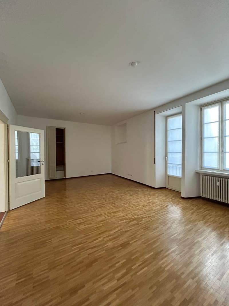Large, open room with hardwood floors, white walls, and large windows. The room appears to be empty, ready for furnishing and decorating.