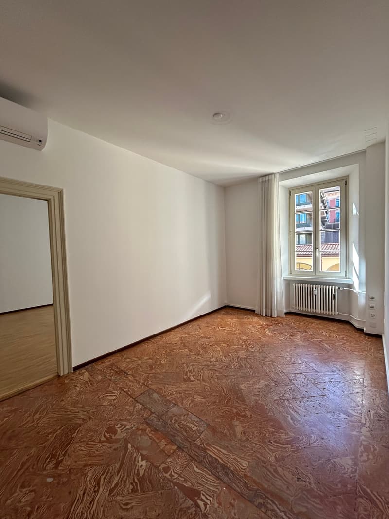 Empty room with white walls, hardwood flooring, and a large window providing natural light. The room appears to be a living room or bedroom with an air conditioning unit installed.