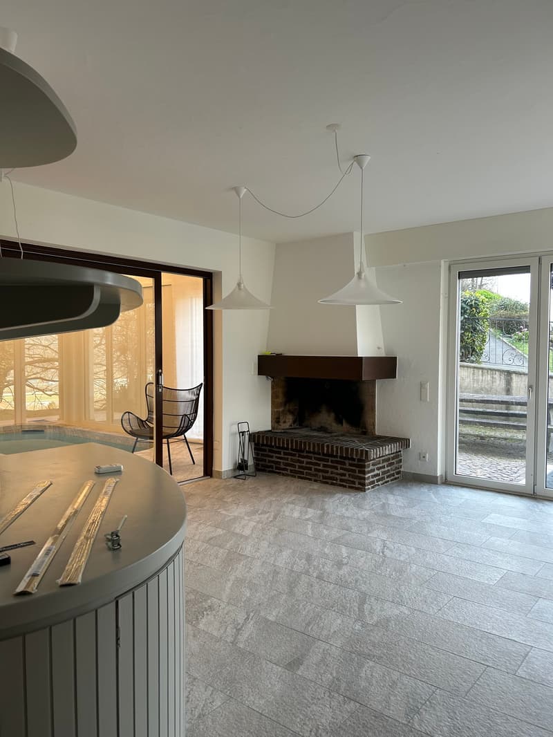 Spacious living room with a brick fireplace, large windows, and modern pendant lighting. The room has a tiled floor and an open layout that connects to other areas of the home.