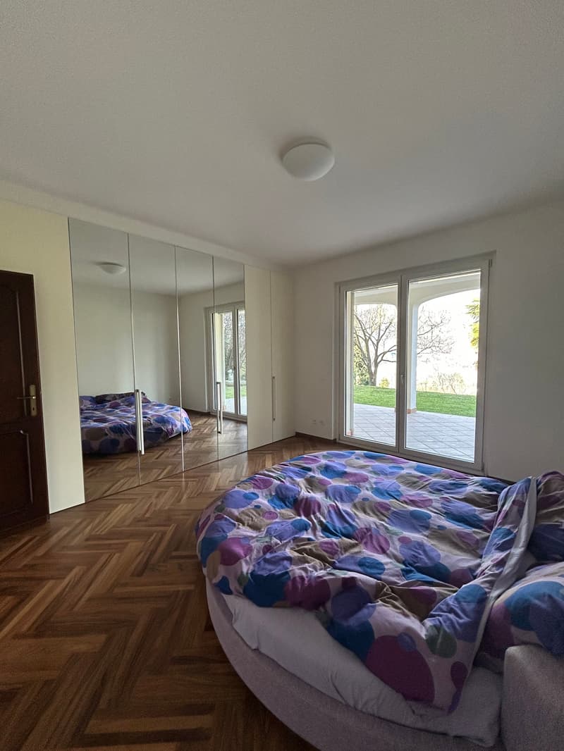 Spacious bedroom with large windows overlooking a garden, hardwood flooring, built-in wardrobe, and a colorful patterned bedding set