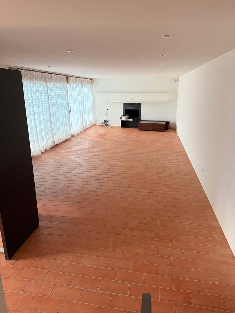 Open room with terracotta tiles flooring, white walls, large white drapes on window, built-in fireplace, TV stand, TV
