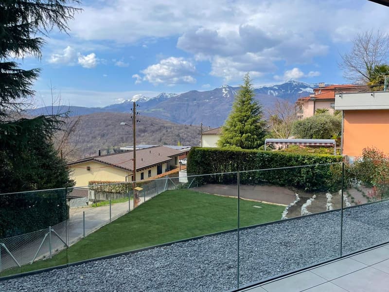 View from a balcony with a glass fence, showing a well-maintained garden with green grass, gravel path, and a hedge. In the background, there are houses, mountains, and a clear sky.