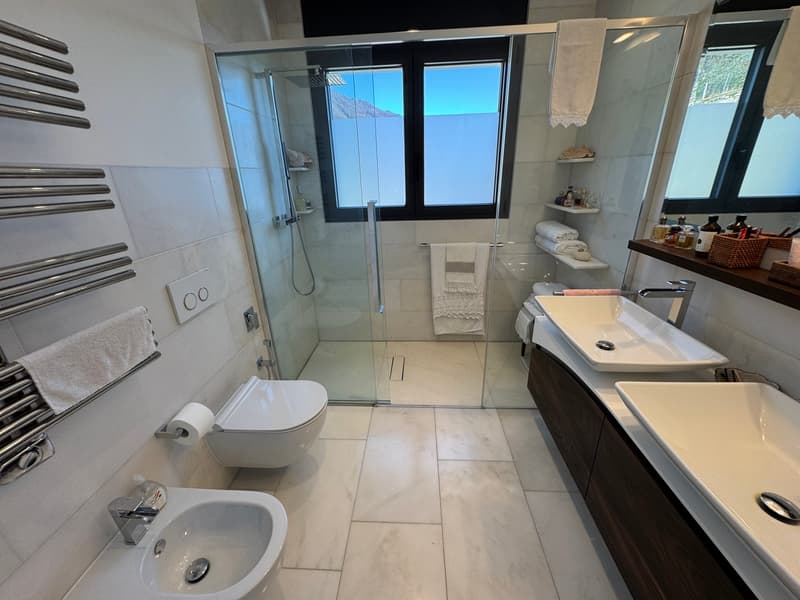 modern bathroom with white tiles, glass shower, two sinks, toilet, windows, towels