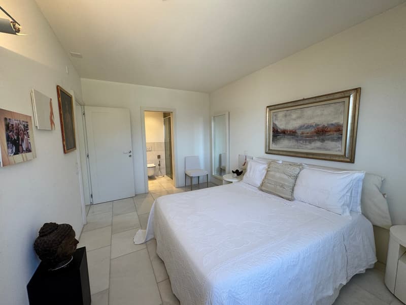 spacious bedroom with a large bed, white walls, white tiled floor, picture frames, chair, bedside tables, Buddha statue, and a painting on the wall