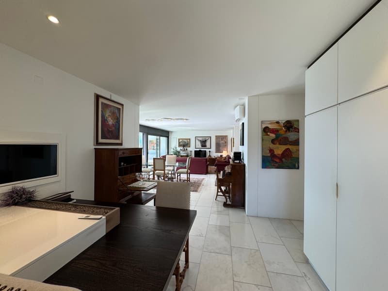 Modern living room with white walls, wooden desk, dining table, cabinets, sofa, TV screen, artwork, tiled floor, and a view outside.