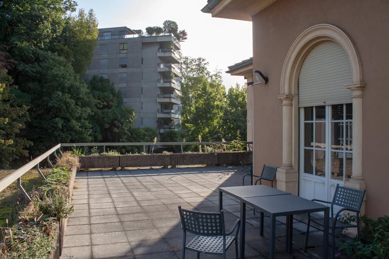 Outdoor area with a table and chairs, surrounded by a fence, with a view of a building and trees.