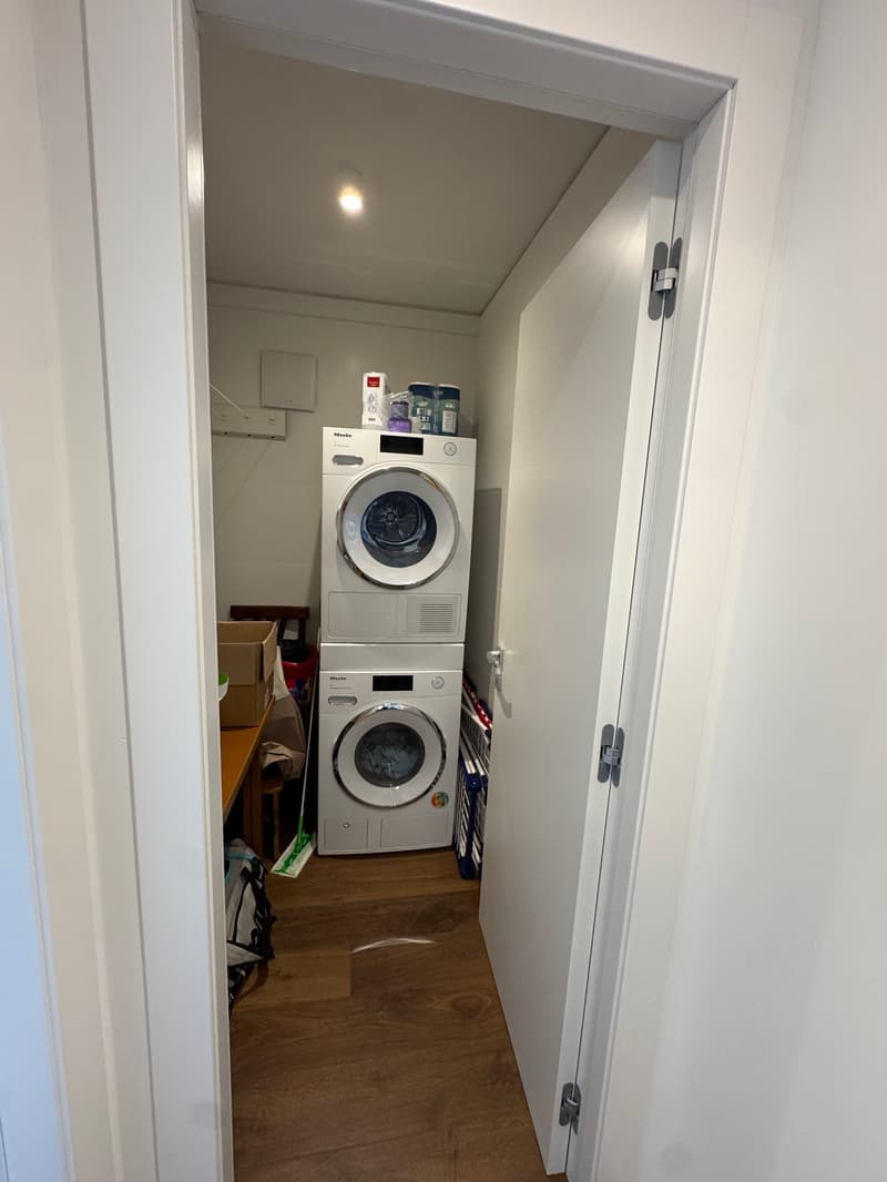A small room with a wooden floor, white walls, and a door. It contains a stack of two washing machines, a table with a box, a red bucket, and a shelf with various items.