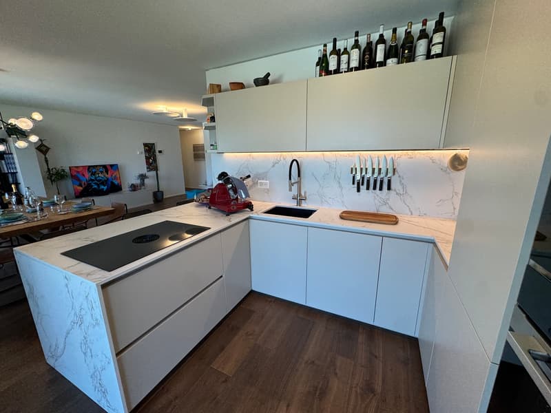 modern kitchen with marble countertops, induction cooktop, wine rack, white cabinets, modern faucet, wine bottles, knives, chopping board, TV, dining table, dining chairs, potted plants