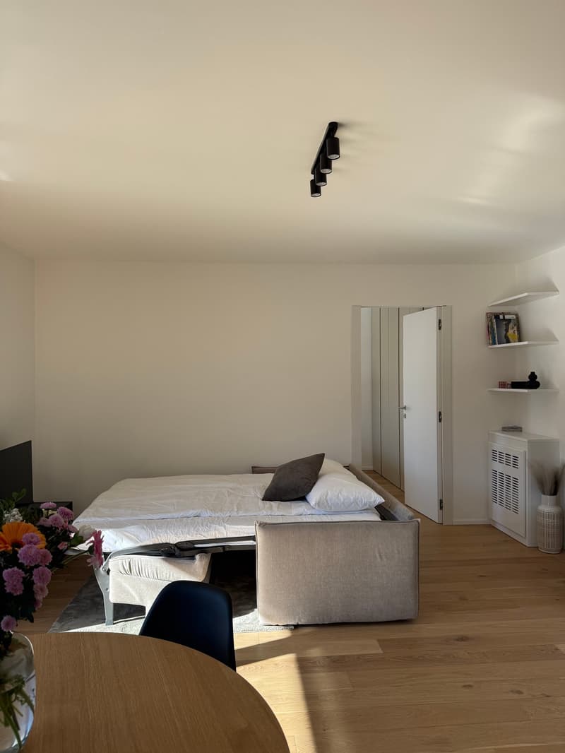 large bed with mattress, side table, chair, wooden floor, white walls, mounted shelves, small television