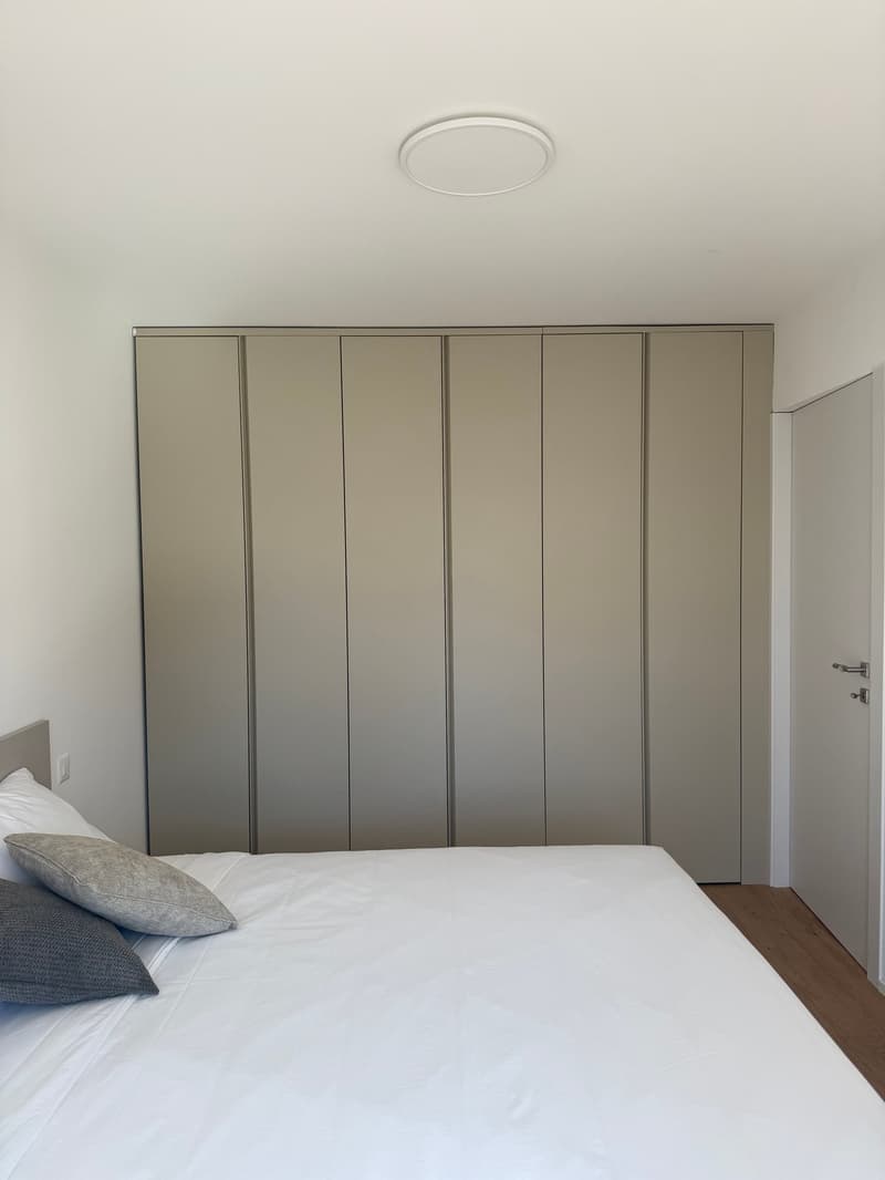 Bedroom with a large bed, white bedding, two pillows, four-door closet, white walls, wooden floor, white ceiling, and a power outlet.