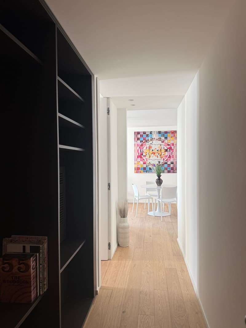 A narrow hallway with wooden flooring, dark cabinet on the left, small table with chairs, a colorful painting, and a white vase with potted plant.