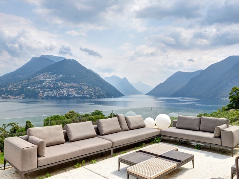 Outdoor seating area with two couches, a coffee table, and a glass railing overlooking a lake and mountains.