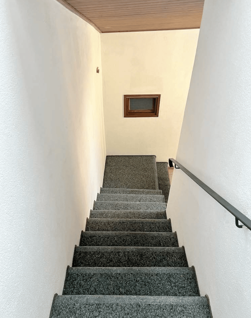 The image shows a staircase with gray granite steps leading up to a wooden ceiling. The walls are painted in a light color, and there is a small window visible on the wall.