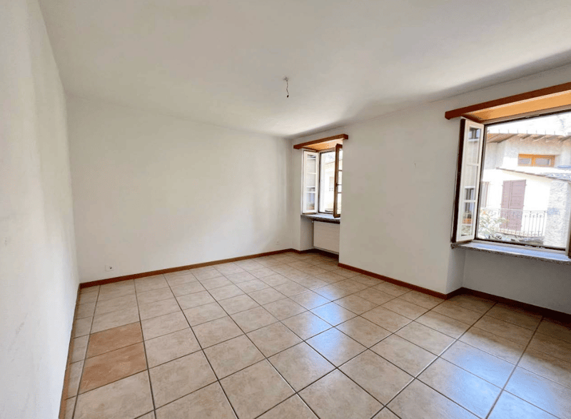 This image shows a spacious room with white walls and a tiled floor. The room has large windows that provide natural light and a view of the outdoors. The room appears to be empty, with no furniture or decor visible.