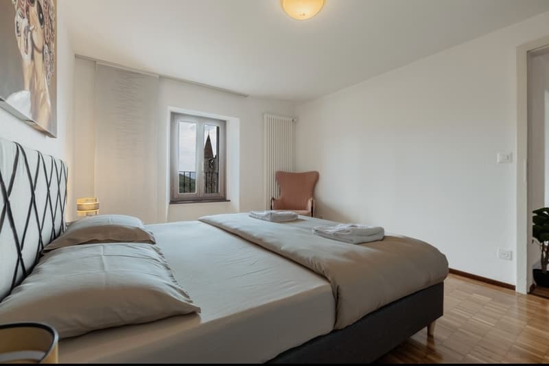Spacious bedroom with hardwood flooring, white walls, large window providing a view, bed with bedding, and an armchair.