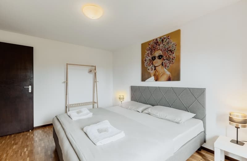 Spacious bedroom with a large bed, gray upholstered headboard, and a colorful abstract painting on the wall. The room has hardwood flooring and a window, suggesting the possibility of a balcony or terrace.