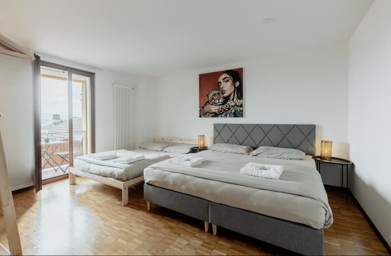 Spacious bedroom with hardwood floors, large windows, two queen-size beds, gray upholstered headboards, side tables, and a piece of artwork on the wall.