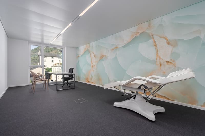 An empty room with a white wall, gray floor, large window, office desk with chair, exam table, and medical chair.