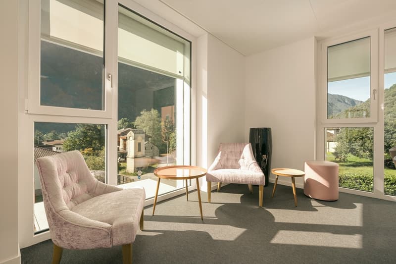 Modern living area with 3 pink chairs, round table, 2 side tables, round pouf, large windows, sun rays