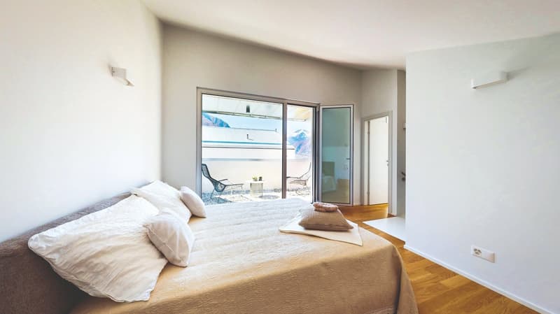 large bed, white pillows, beige blanket, balcony with chairs, table, scenic view