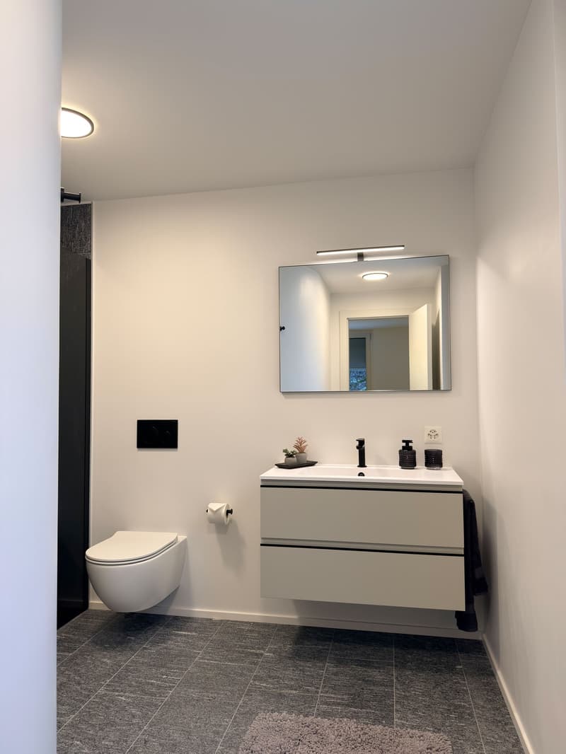 modern bathroom, with white tile, white vanity, mirror, toilet, grey tile flooring, white ceiling, wall outlet, roll of toilet paper