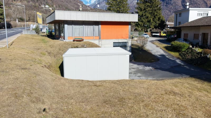 A modern building, painted in a combination of white and orange, nestled against the base of a mountain. The driveway to the house is made of asphalt and leads to an underground garage. To the left of the house, there is a road with street signs and traff