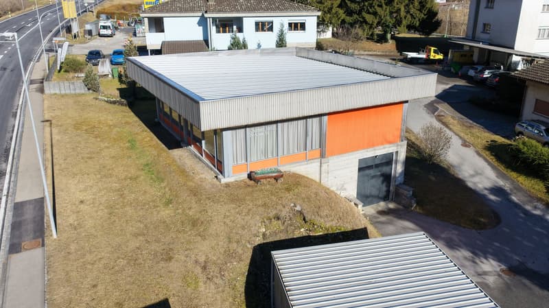Industrial building with gray and orange panels, metal roof, garage entrance, surrounded by grassy terrain, multiple parked vehicles, nearby roadway, other buildings