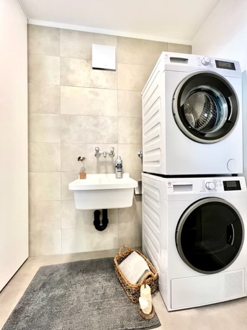 Two white stackable washing machines, a small white sink, a basket on the floor