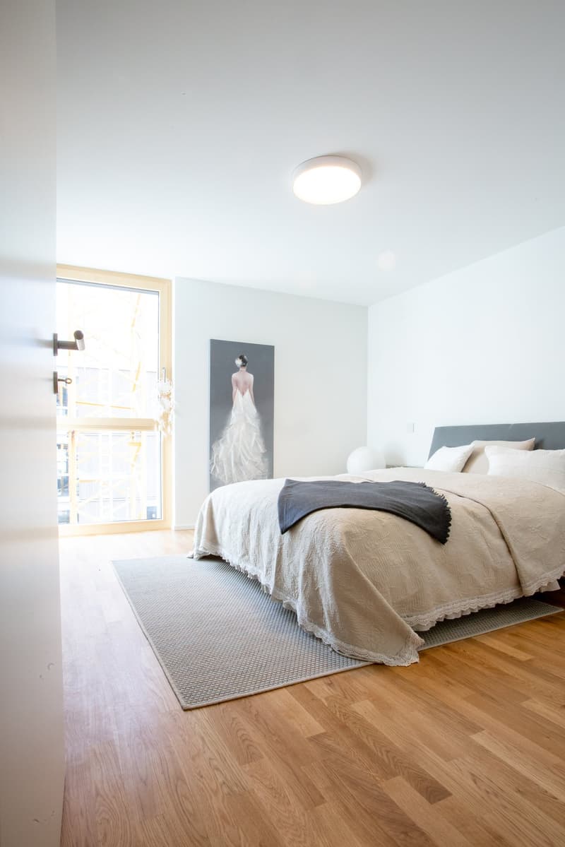 wooden floor, large double bed, white duvet, gray rug, white door, framed painting on the wall, round glass door to balcony