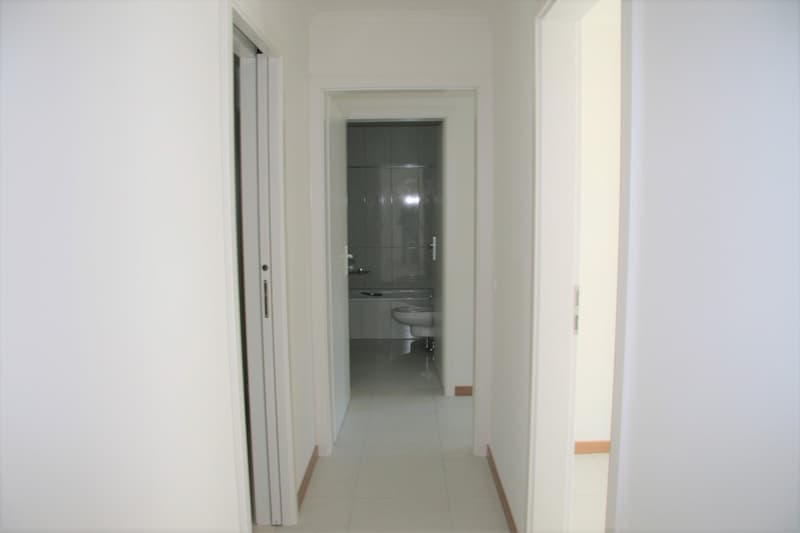Empty hallway with white walls and tiled floor, leading to a bathroom with a toilet and bathtub.