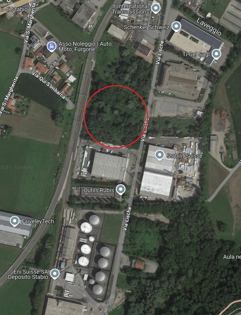 Industrial area with several buildings, open spaces, and trees, roads and streets, green field in the background