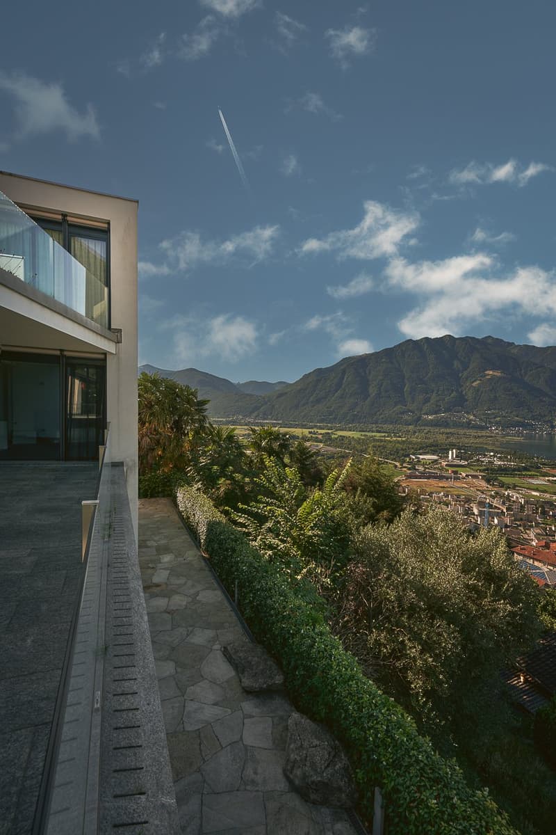 Contemporary house with glass balcony, stone walkway, lush greenery, distant mountain range, and cityscape view