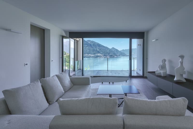 open living space, large grey sofa, glass coffee table, large glass windows, balcony, lake and mountain view