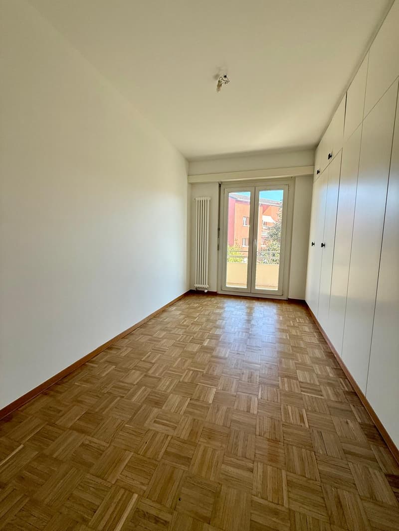 empty room, wooden parquet flooring, white walls, large windows, sliding glass door to balcony, built-in white cabinets