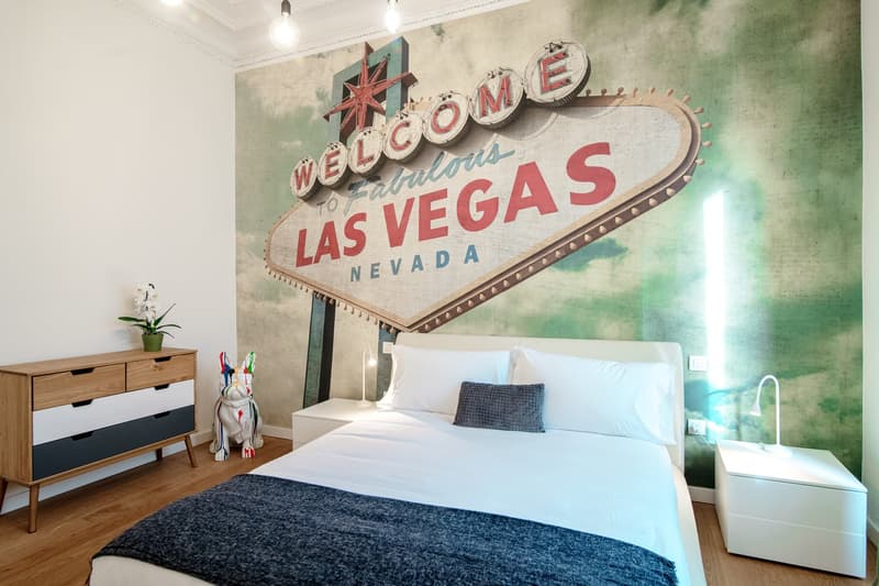 Bedroom with Las Vegas sign mural, white bedding, lamp, nightstand, chest of drawers, statue