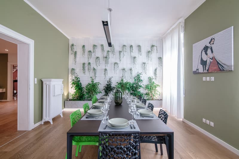Dining room with a large table and green chairs, white radiator, plants in the background, white ceiling with pendant lighting