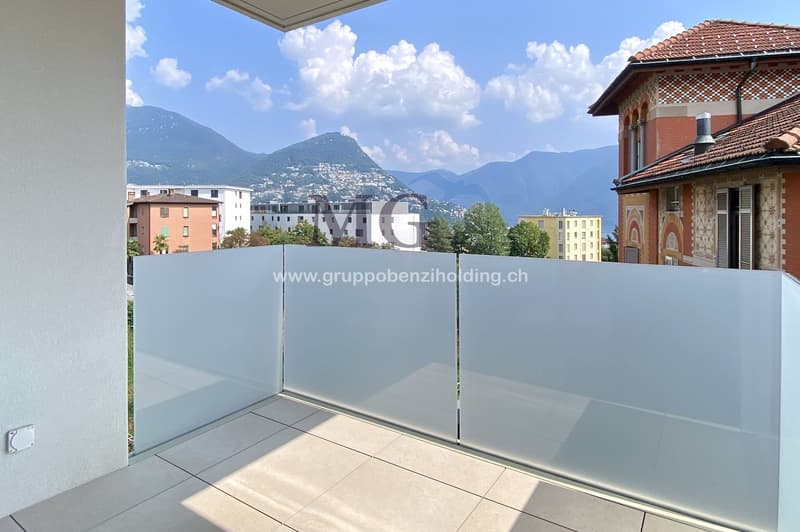 A balcony with white railing and glass partition offering a scenic view of mountains and cityscape.
