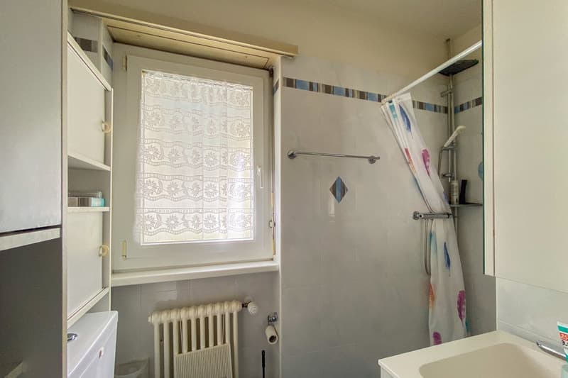 tiled walls, white window with lace curtain, white sink, white toilet, white radiator, shower with shower curtain