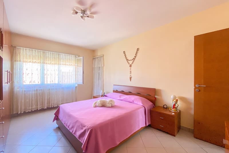 a bedroom with a wooden bed, pink bedding, wooden nightstand, white wall, tiled floor, and window with curtains