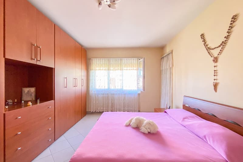 Bedroom with double bed, wooden wardrobes, a framed photo, a window with curtains, a wooden nightstand with items on top, and a rosary hanging on the wall.