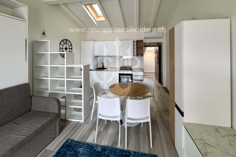 A modern, minimalistic dining area with a wooden table and white chairs. The room is furnished with white shelves, a gray sofa, and features wooden floors and white walls. A kitchen with a modern sink, a gas stove, and a refrigerator is also visible.